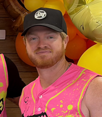 Man with red hair and beard wearing a black cap and a bright pink shirt.