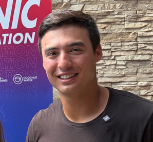 Young man smiling, wearing a dark shirt, with a stone wall background.