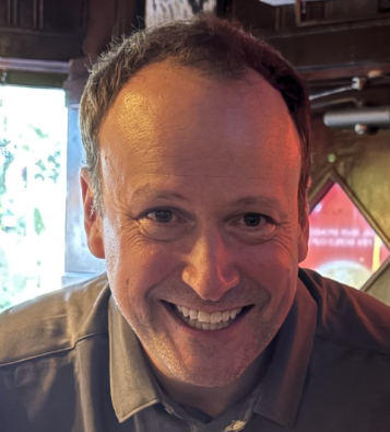 Smiling man with short hair, wearing a collared shirt, in a warmly lit indoor setting.