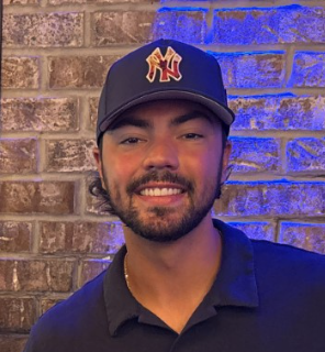 Young man with a beard wearing a baseball cap, smiling in front of a brick wall.