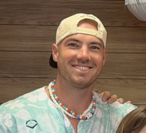 Smiling man in a floral shirt and baseball cap, wearing a beaded necklace.