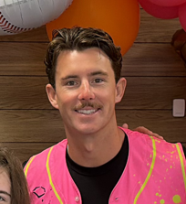 Young man wearing a pink vest, smiling against a backdrop of colorful balloons.