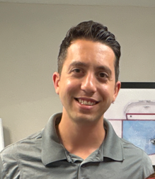Smiling man wearing a gray shirt, standing in an office setting.